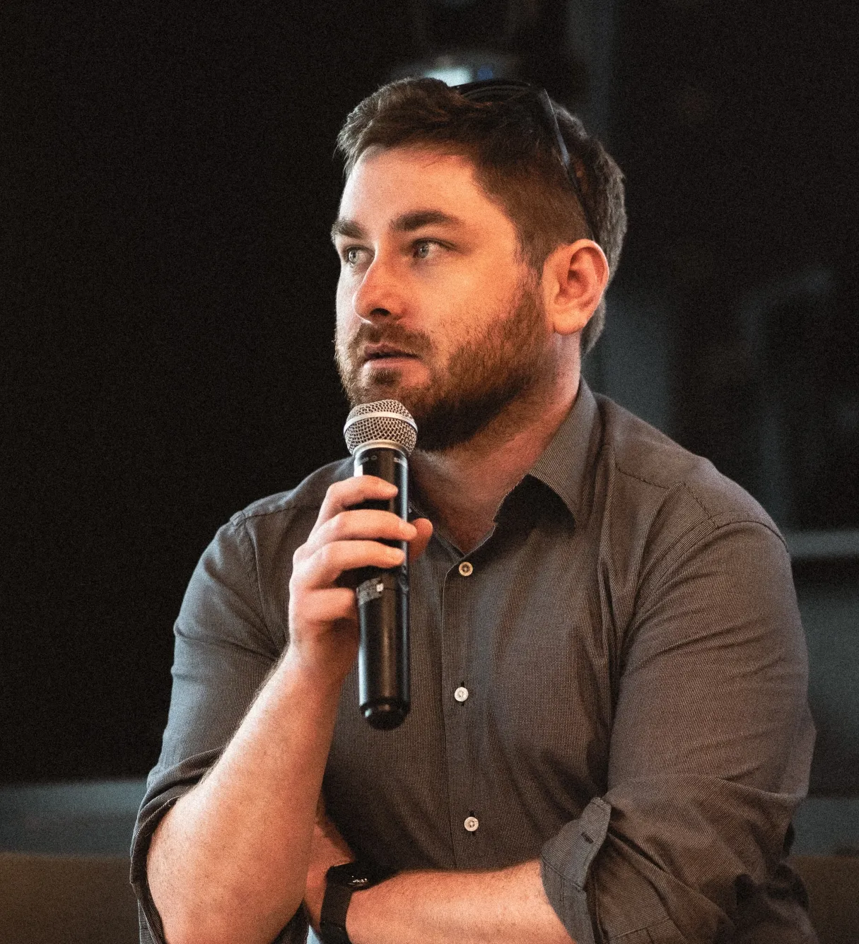 Callum holding a microphone at a speaking event, wearing a dark shirt with sunglasses resting on his head.